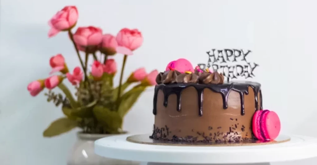 Eggless chocolate birthday cake with chocolate drip, pink macarons, and Happy Birthday topper on white cake stand with pink tulips