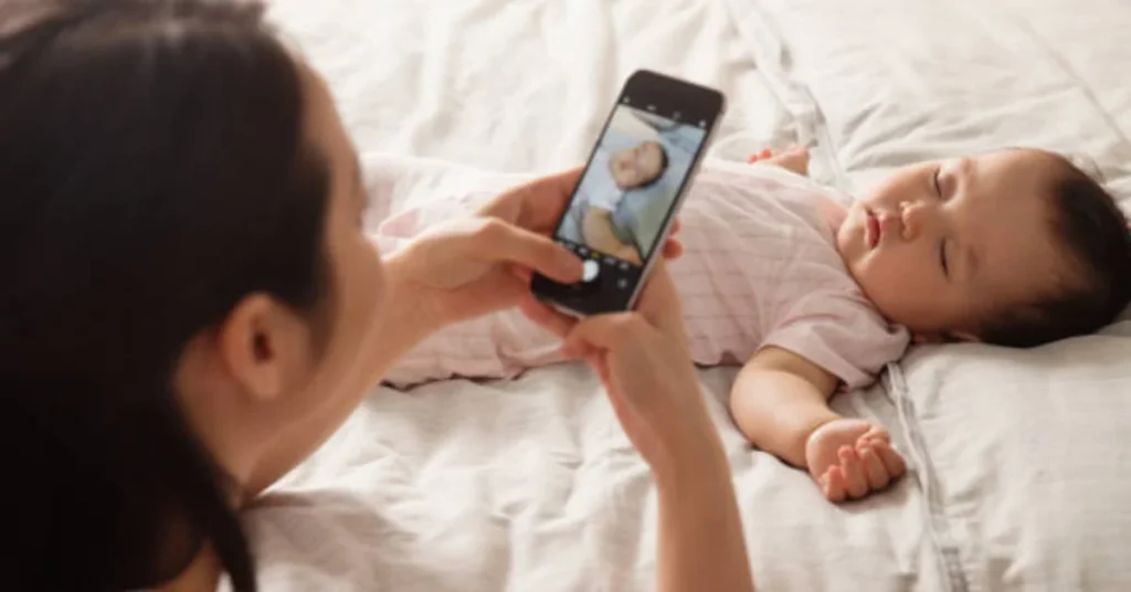 Mother taking a photo of her peacefully sleeping 9-month-old baby on white bedding with smartphone