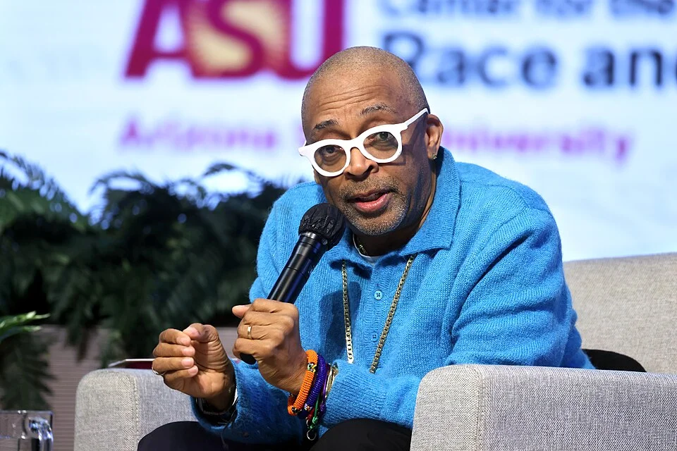 Spike Lee speaking at ASU event wearing signature white glasses and blue sweater