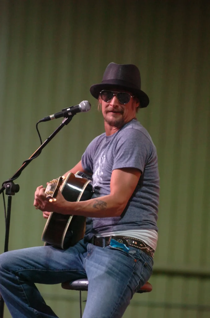 Kid Rock performing acoustic guitar on stage wearing hat and sunglasses