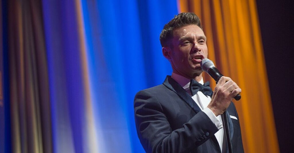 Ryan Seacrest hosting event in tuxedo with microphone