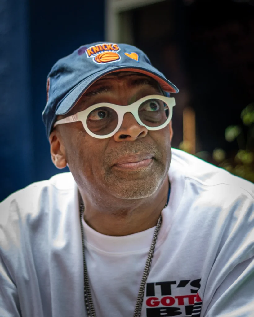 Spike Lee wearing Knicks cap and signature white glasses in casual portrait