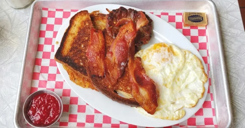 A diner breakfast platter on a metal tray with fried eggs, bacon, sausage, toast, hash browns, jam, and butter.