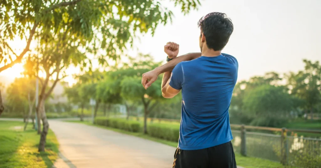 Person in blue shirt stretching arm outdoors on park path during early morning golden hour sunlight