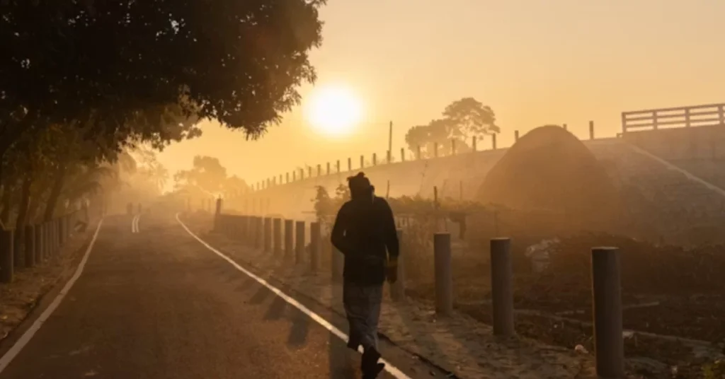 Person walking along a quiet road at sunrise, with golden light, trees, and misty countryside in the background.