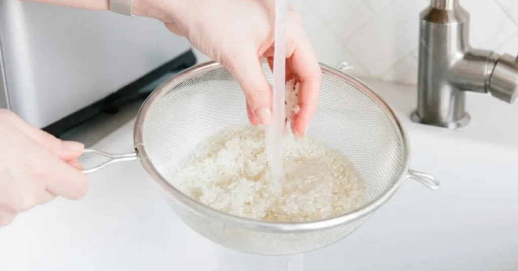 A close-up shot of hands rinsing white rice in a metal mesh strainer under a running stream of tap water in a white kitchen sink.