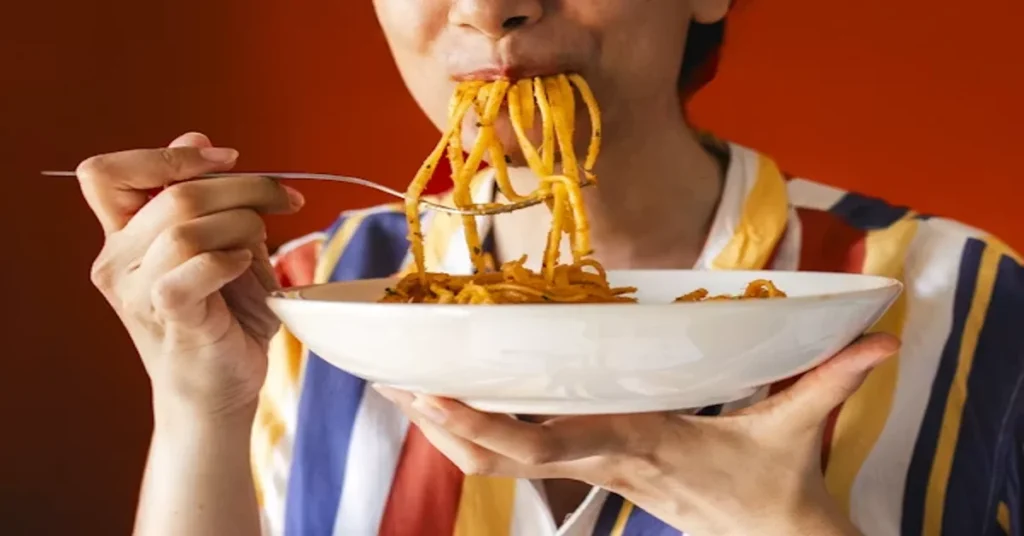 Person eating spaghetti from a white bowl against a red background.