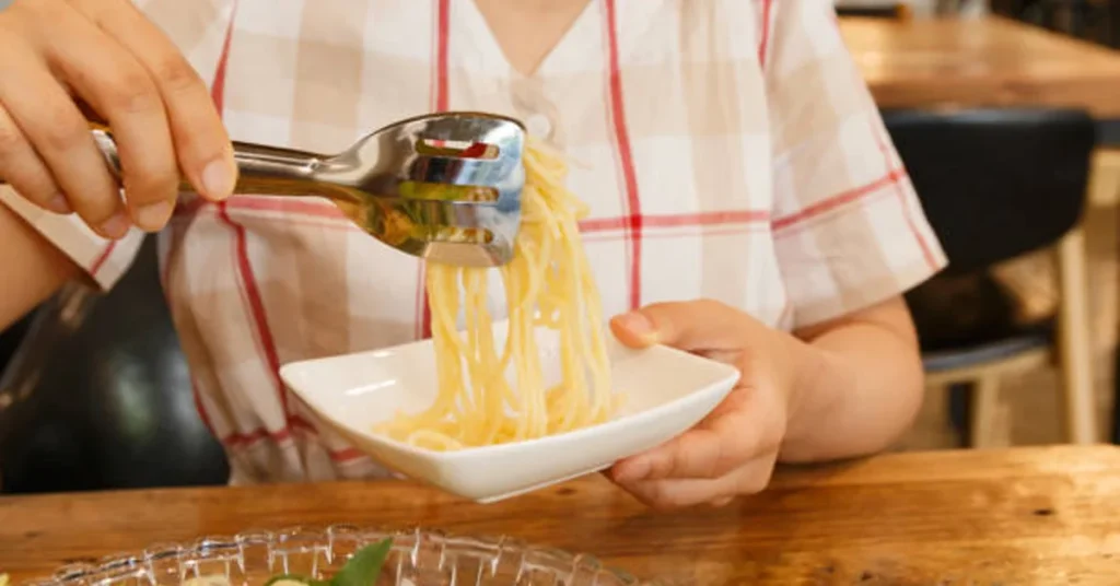 Person serving cooked spaghetti into a small bowl with pasta tongs at a wooden table.