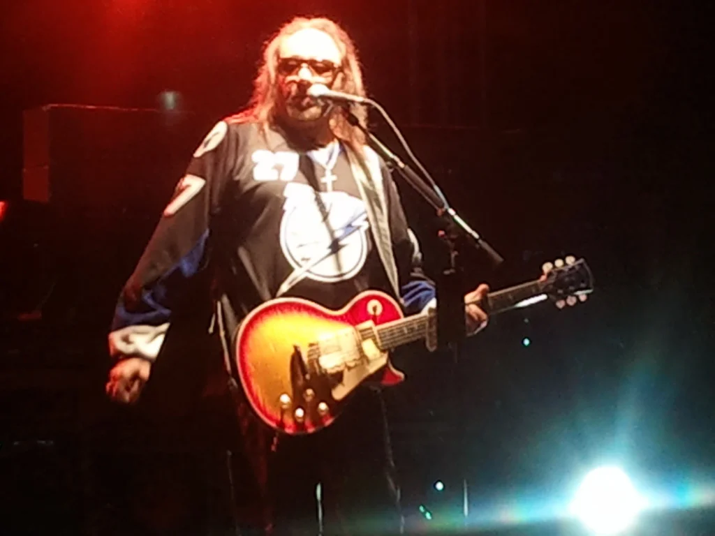 Ace Frehley performing on stage with sunburst Les Paul guitar wearing hockey jersey and sunglasses