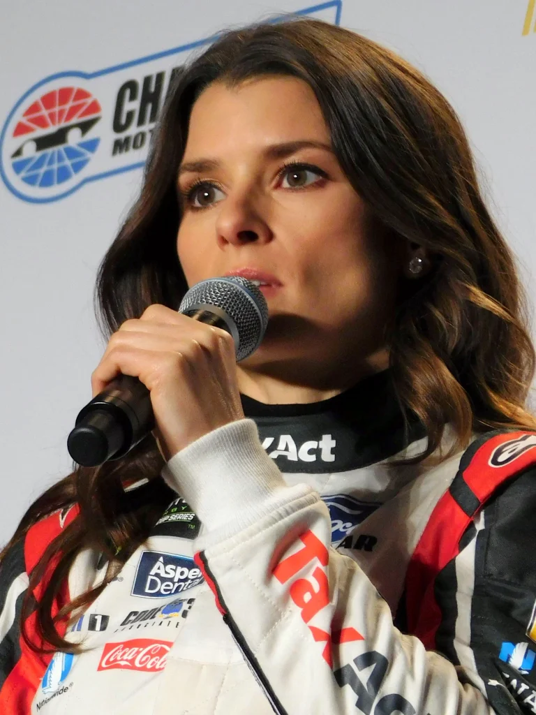 Danica Patrick at NASCAR press conference wearing racing suit with sponsor logos, speaking into microphone