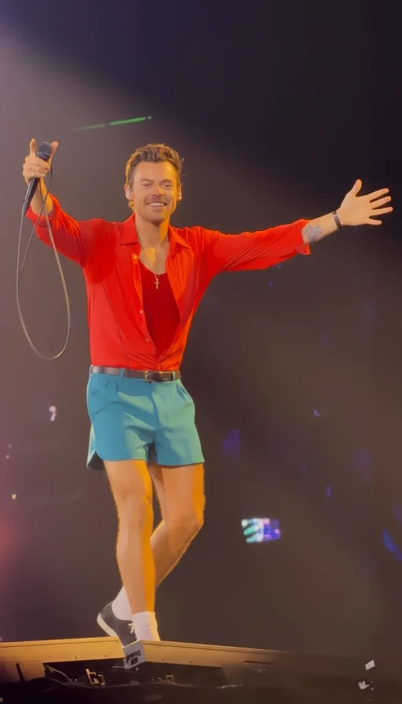 Harry Styles performing on stage in red shirt and blue shorts with arms outstretched